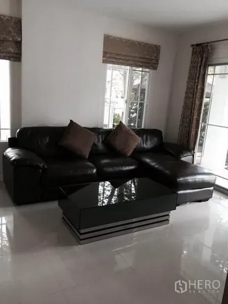 Detached House for rent in Bang Kapi, Bangkok - Living room with black leather sectional sofa and low coffee table beside large windows.