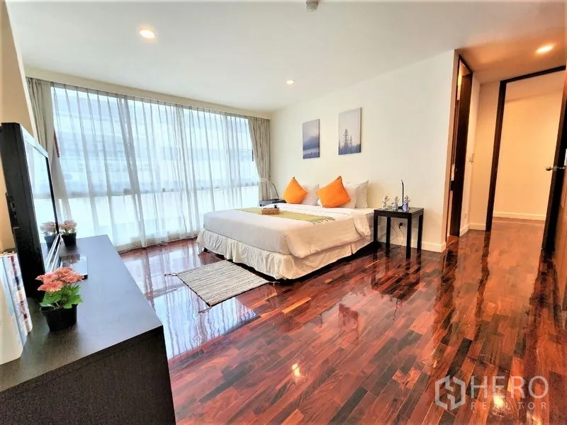 Condominium for rent in Bang Rak, Bangkok - Spacious bedroom with TV stand, wide windows and warm wood floors accented with orange cushions.
