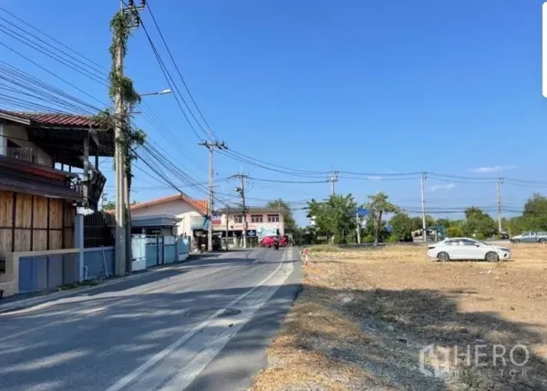 Land for sale in Mueang Samut Prakan, Samut Prakan - Corner of a local paved road beside a cleared land plot in Phraeksa with power lines and houses under a blue sky