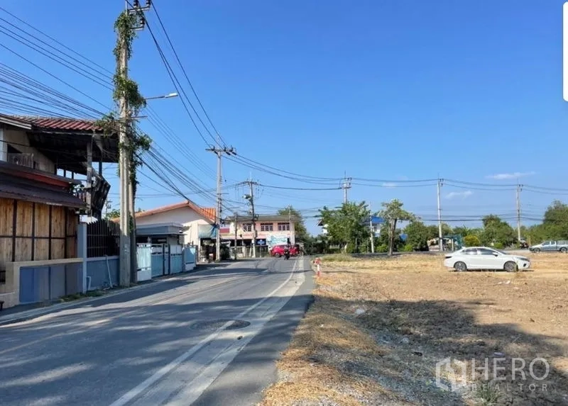 Land for sale in Mueang Samut Prakan, Samut Prakan - Corner of a local paved road beside a cleared land plot in Phraeksa with power lines and houses under a blue sky