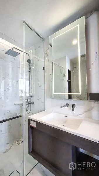 Condominium for sale in Khlong Toei, Bangkok - Modern bathroom featuring marble-pattern walls, glass shower with rain head, and backlit mirror above sink.