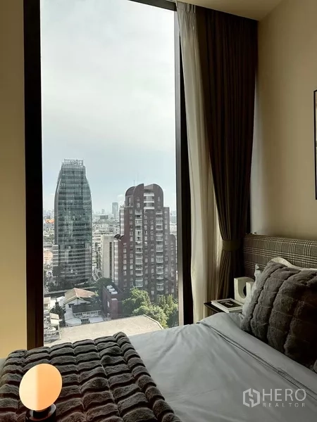 Condominium for rent in Pathum Wan, Bangkok - Second bedroom with a tall window and urban view, cozy bedding and a nightstand.