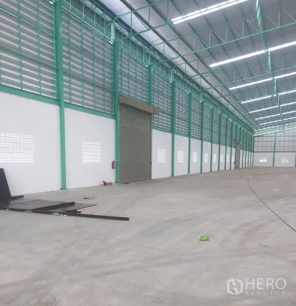 Warehouse for rent in Samut Prakan - Long warehouse bay with louvered ventilation, wide roller shutter door, and smooth concrete floor under a metal roof.
