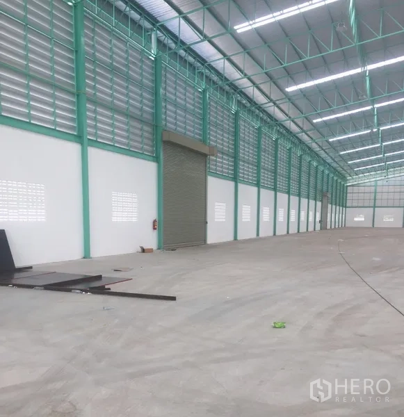 Warehouse for rent in Samut Prakan - Long warehouse bay with louvered ventilation, wide roller shutter door, and smooth concrete floor under a metal roof.