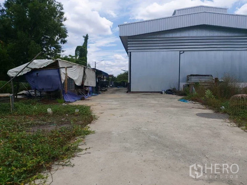 Warehouse for sale in Mueang Nonthaburi, Nonthaburi - Wide concrete driveway beside a large metal-clad warehouse with tarp-covered sheds and greenery.