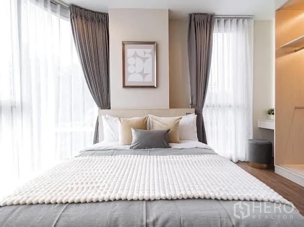 Condominium for rent in Watthana, Bangkok - Cozy queen bed framed by dual corner windows and soft gray curtains.