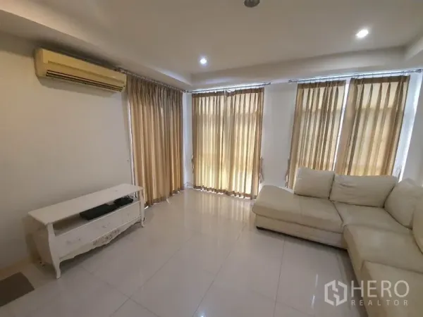 Townhouse for rent in Phaya Thai, Bangkok - Bright living room with beige curtains, recessed lights, split A/C and an L-shaped sofa on glossy tiled floors.