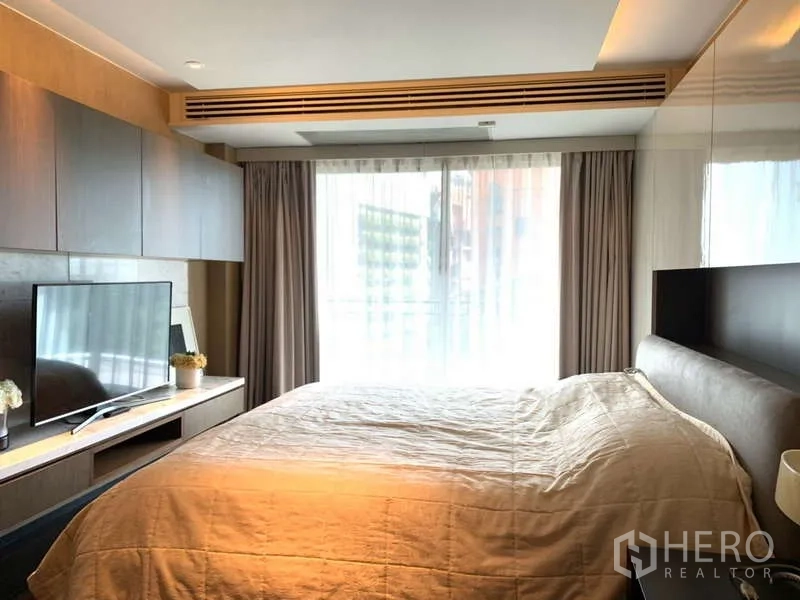 Condominium for rent in Khlong Toei, Bangkok - Sunlit bedroom with built-in storage, TV and floor-to-ceiling window drapes.