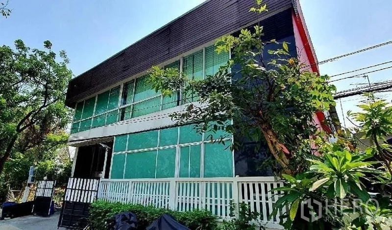 独立商业地产 出租或出售在Bang Khun Thian, Bangkok - 两层独栋建筑，绿色玻璃立面，被茂密树木环绕。