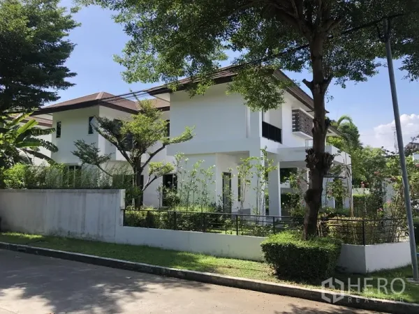 Detached House for rent in Saphan Sung, Bangkok - Side view of a white two-storey home surrounded by mature trees and a low boundary wall.