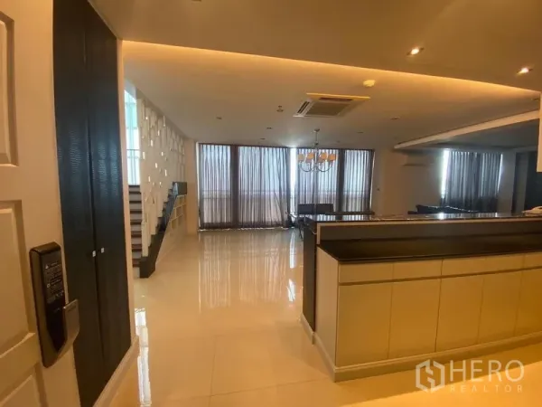Condominium for rent in Bang Kapi, Bangkok - Entry to a spacious living–dining area with glossy floors, kitchen counter and staircase beside tall curtained windows.