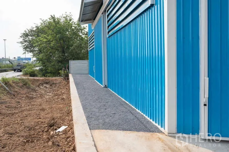 货栈 出租在Khlong Luang, Pathum Thani - 仓库侧立面，沿墙碎石步道，旁临道路与树木。