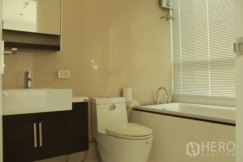 Condominium for rent or sale in Watthana, Bangkok - Second bathroom with vanity, toilet, and bathtub by a window with blinds.