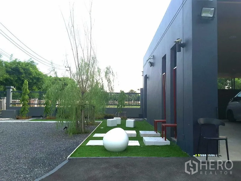 บ้าน ให้เช่าหรือขายในLam Luk Ka, Pathum Thani - สวนข้างบ้านปูหญ้า มีทางเดินแผ่นทางเท้า เก้าอี้ตกแต่ง และต้นไม้เรียงข้างกำแพงสีเทา