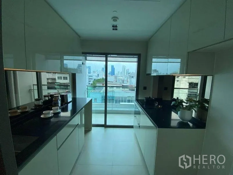 公寓 出售在Bang Rak, Bangkok - 现代一字型厨房，白色亮面橱柜与黑色台面，推拉门连通阳台与城市景观。