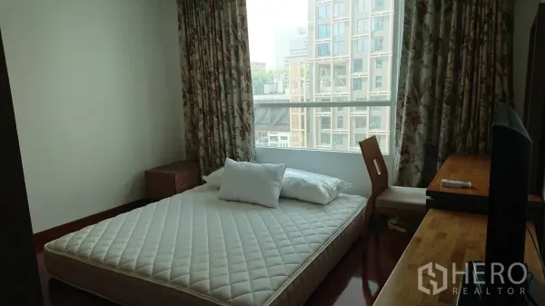 Condominium for rent in Pathum Wan, Bangkok - Bedroom with large window, floral curtains, a low bed frame with mattress and a wooden desk.