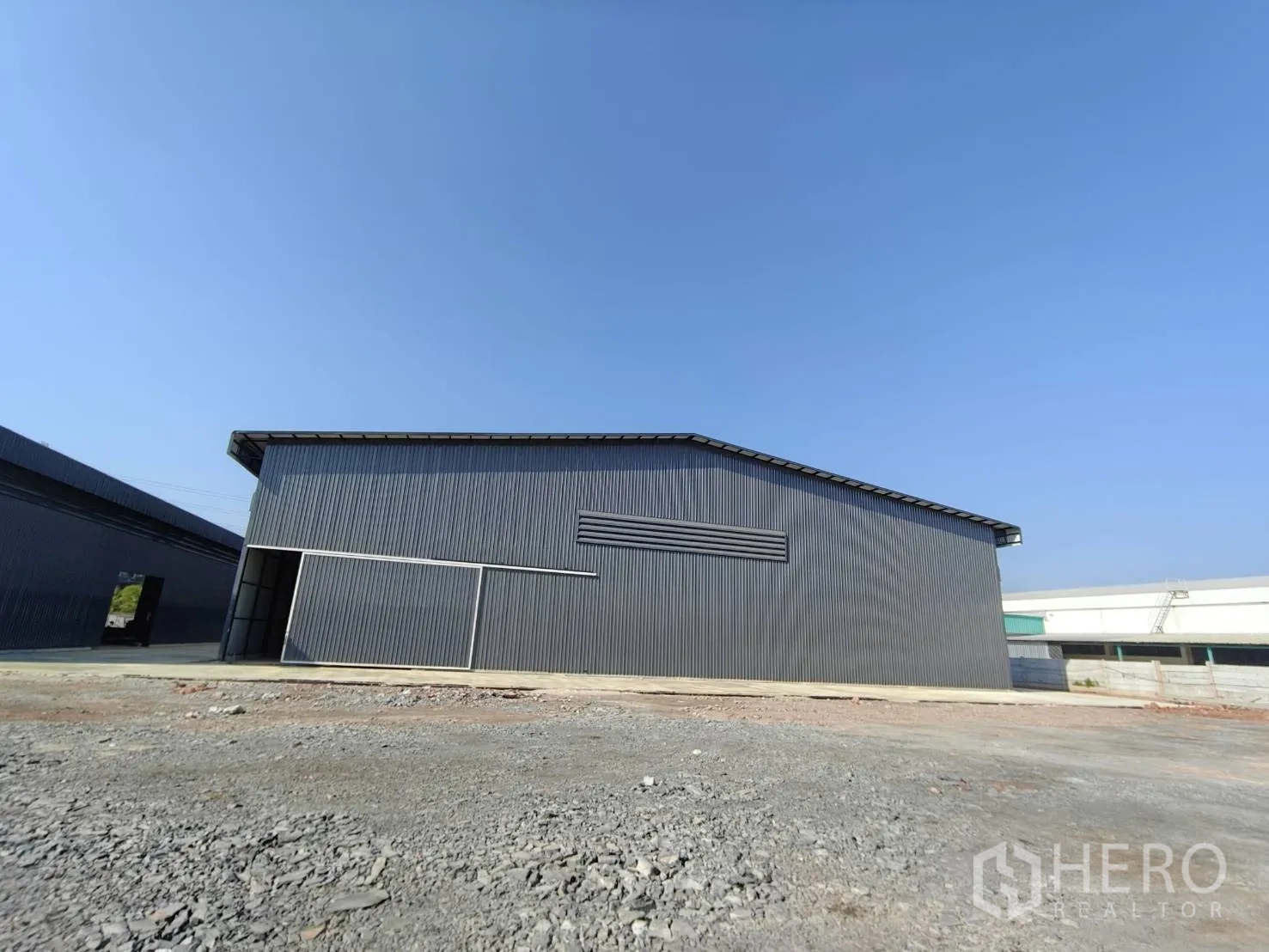 Factory for rent in Mueang Samut Sakhon, Samut Sakhon - Side view of a corrugated steel factory building with an open sliding door and gravel forecourt under blue sky.