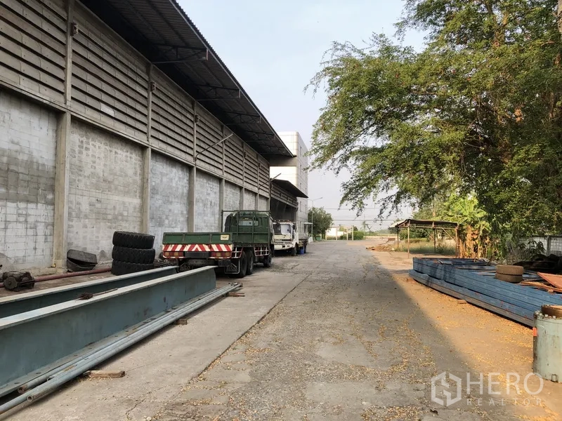 工厂 出租在Bang Bua Thong, Nonthaburi - 厂房侧院，有卡车、钢梁与成排大树