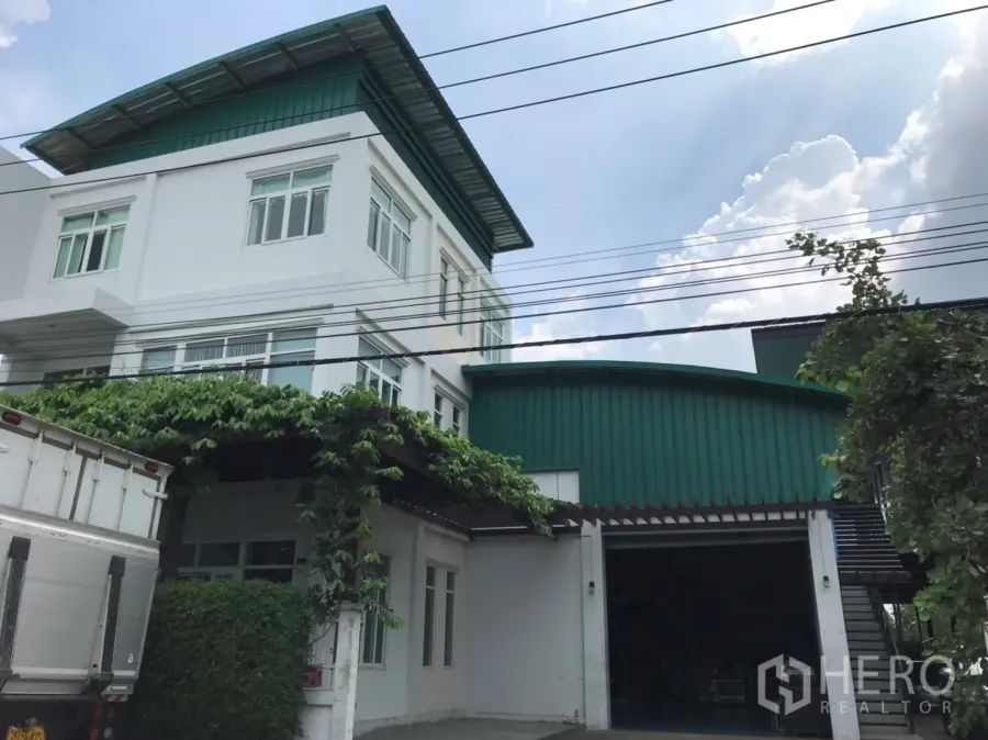 工厂 出售在Lam Luk Ka, Pathum Thani - 绿色金属屋顶与白色办公楼相连的工厂外观，设宽卷闸与车道