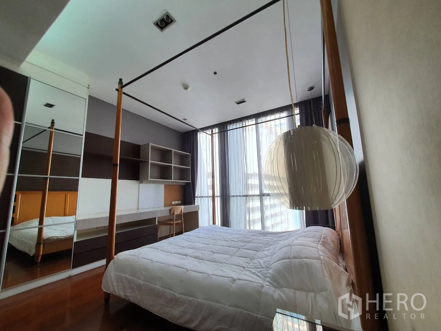 Condominium for rent in Khlong Toei, Bangkok - Bedroom with four-poster bed, working desk, shelving and mirrored wardrobe by tall windows.