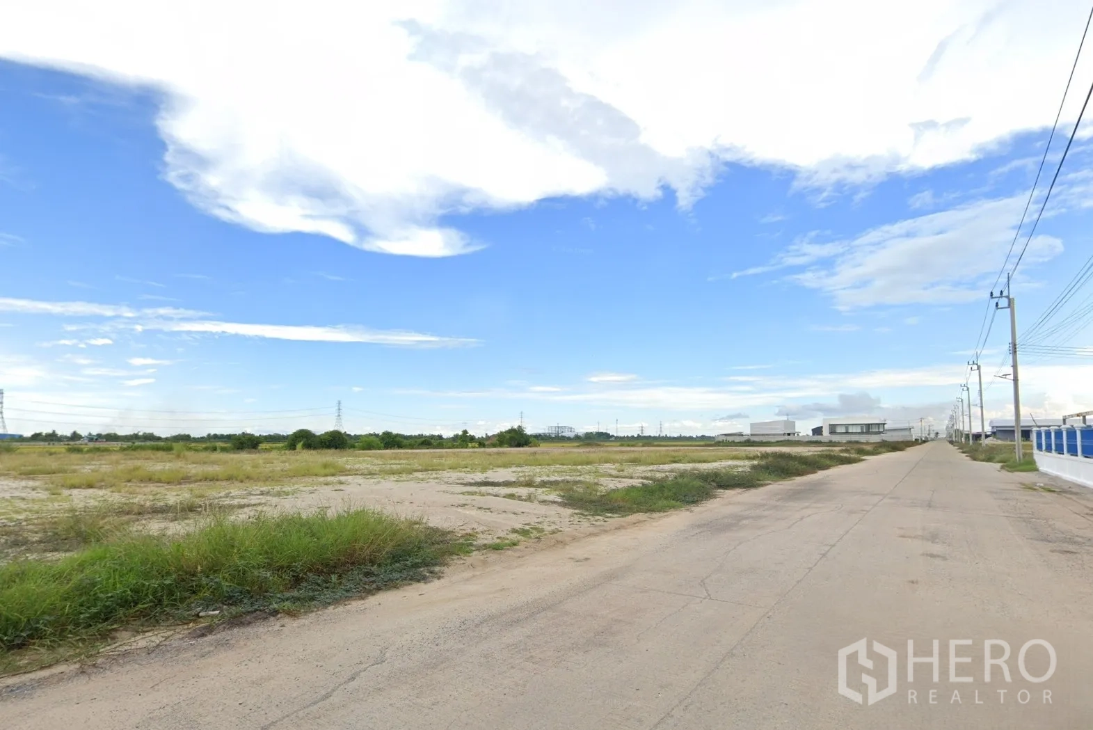 Land for sale in Sai Noi, Nonthaburi - Roadside view showing the plot, utility poles, and nearby warehouse buildings.