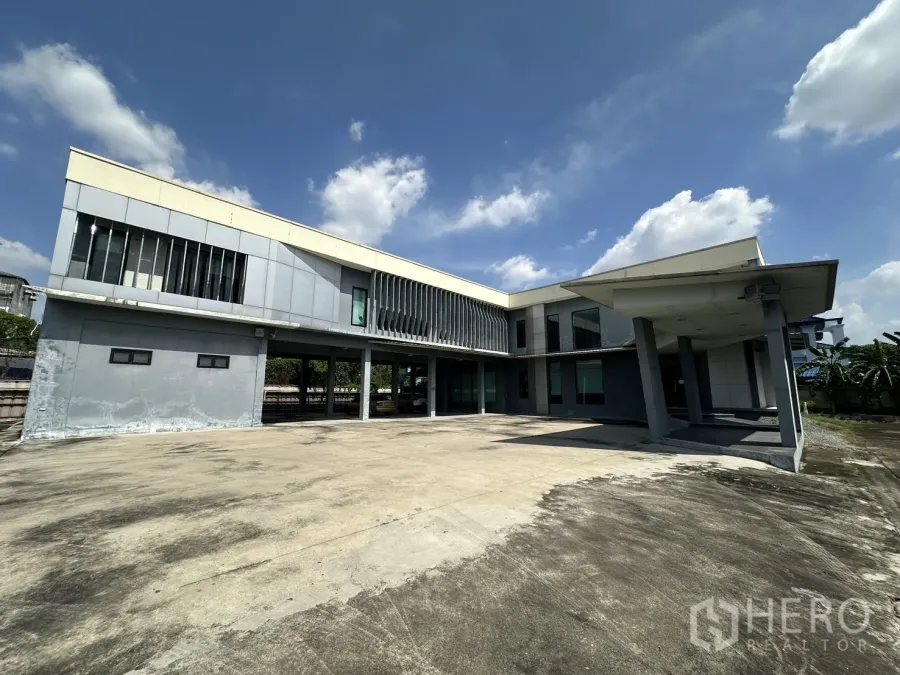 Home Office for rent in Prawet, Bangkok - Front facade of the two-storey office building with expansive parking apron under a blue sky.