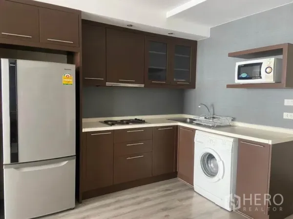 Condominium for rent in Watthana, Bangkok - L-shaped kitchen with brown cabinets, cooktop, fridge, microwave, and washing machine.