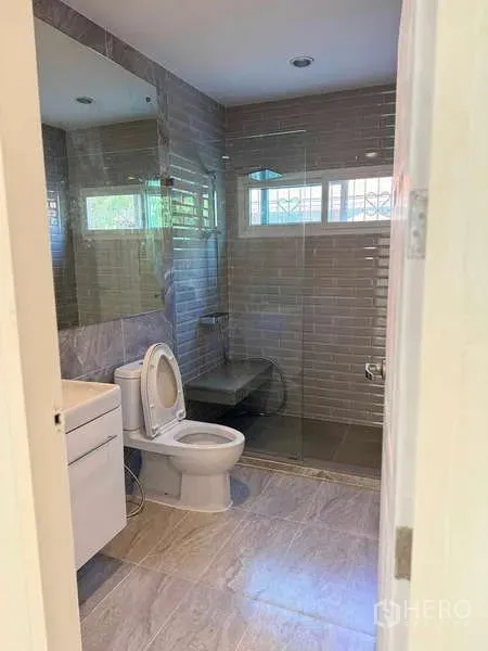 Detached House for rent in Samut Prakan - Modern bathroom with glass-enclosed shower, gray wall tiles, vanity, and toilet.