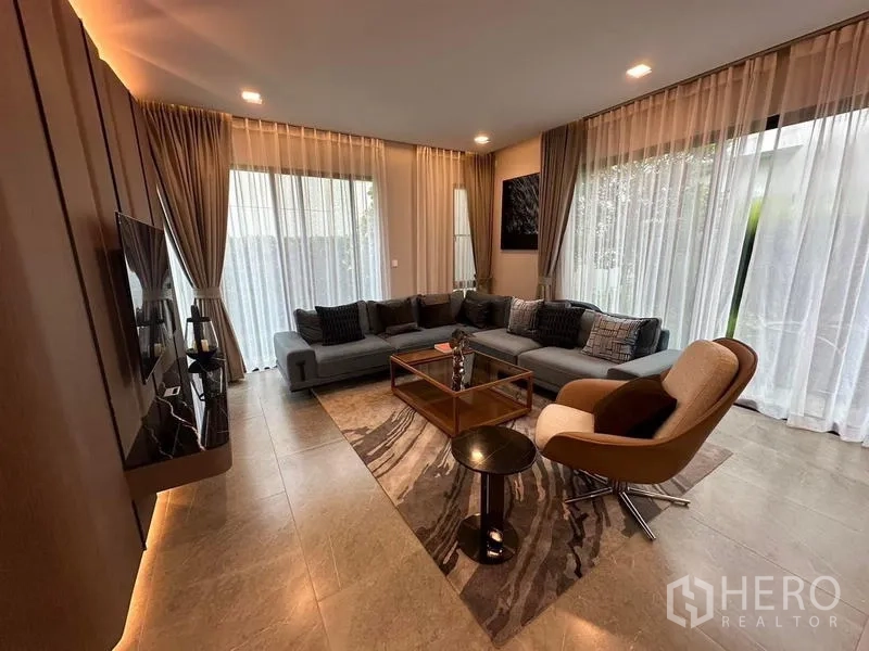Detached House for rent in Samut Prakan - Living room with L-shaped sofa, armchair and sheer curtains.