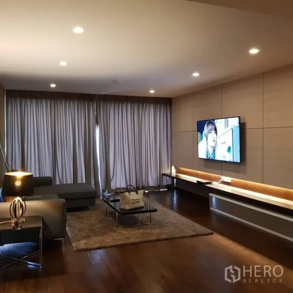 คอนโด ให้เช่าหรือขายในPathum Wan, Bangkok - ห้องนั่งเล่นอบอุ่น โซฟาเข้ามุม ทีวีติดผนัง ไฟดาวน์ไลต์ และผ้าม่านเต็มความสูง