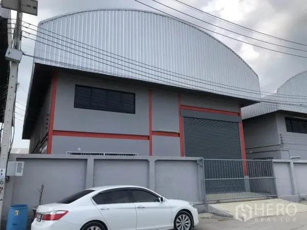 货栈 出租在Nonthaburi - 灰红配色的仓库外观，弧形金属屋顶、大型卷闸门和带门禁入口，临混凝土道路。