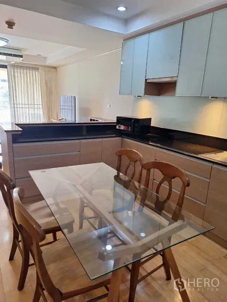 Apartment for rent in Watthana, Bangkok - Glass dining table with wooden chairs beside a light-wood kitchen with black countertops and upper cabinets.