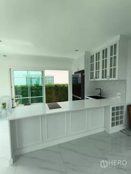 Detached House for rent in Bang Phli, Samut Prakan - White kitchen with long peninsula, glass-front cabinets, and sliding doors opening to the garden.