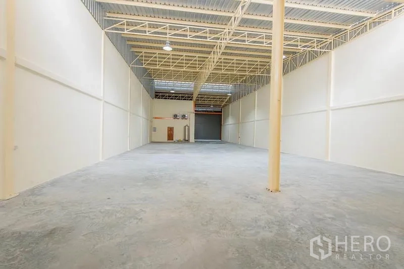 Factory for sale in Bang Sao Thong, Samut Prakan - Spacious warehouse interior with high steel-truss ceiling, skylights and smooth concrete floor leading to a roller shutter.