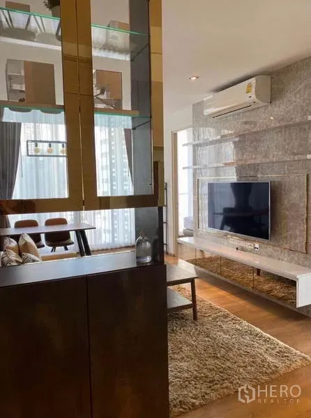 Condominium for rent in Khlong Toei, Bangkok - Entry display cabinet with glass shelves overlooking lounge with mounted TV and tall windows.