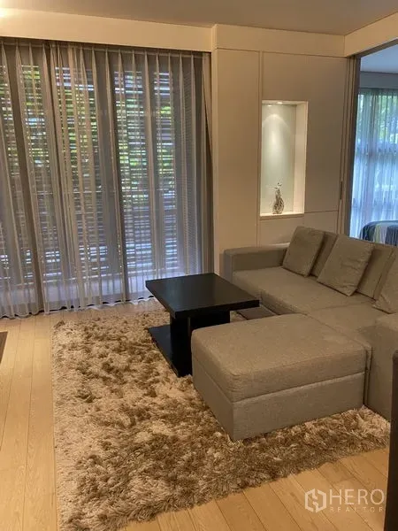 Condominium for rent in Watthana, Bangkok - Bright living room with a gray sectional sofa, shag rug, and floor‑to‑ceiling sheer curtains.