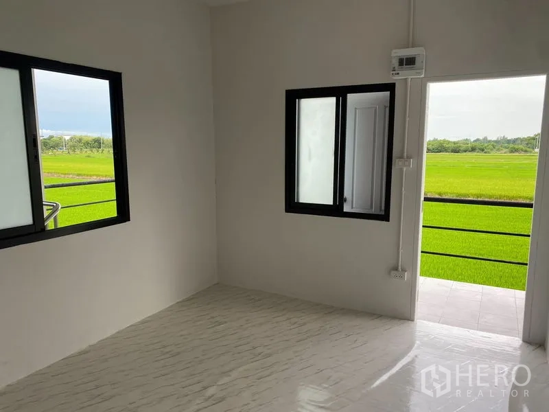 Warehouse for rent or sale in Bang Len, Nakhon Pathom - Compact room with windows and a balcony door facing green fields.