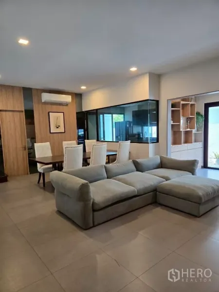 Townhouse for rent in Bang Phli, Samut Prakan - Open-plan living and dining room with large grey sectional sofa, dining table, and a glass-partitioned kitchen in a modern townhouse.