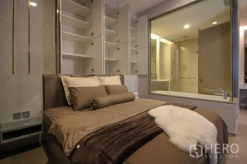 Condominium for sale in Watthana, Bangkok - Bedroom angle showing built-in shelving and a glass partition to the bathroom under warm ambient lighting.