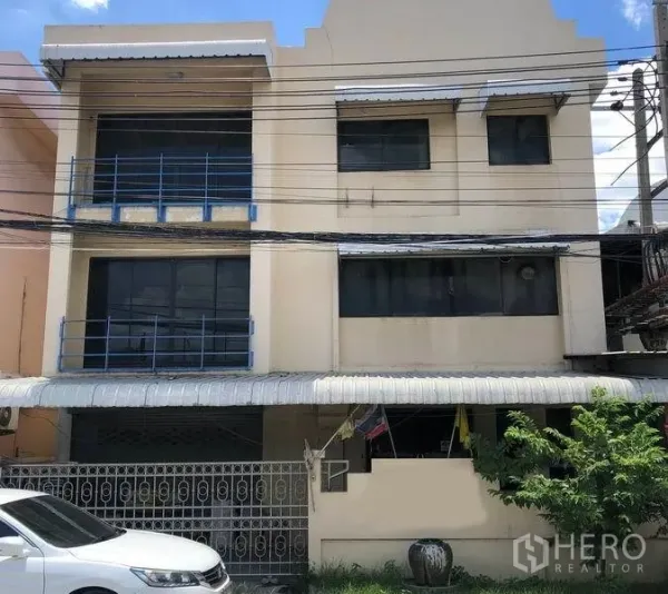 工厂 出售在Bang Bua Thong, Nonthaburi - 暖米色三层工厂外立面，蓝色栏杆，带大门与遮棚的前院车位，位于暖武里府邦武通。