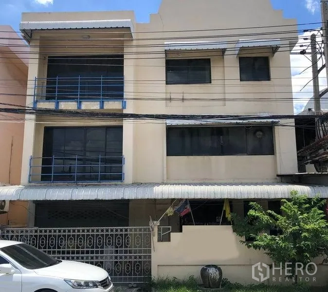 工厂 出售在Bang Bua Thong, Nonthaburi - 暖米色三层工厂外立面，蓝色栏杆，带大门与遮棚的前院车位，位于暖武里府邦武通。