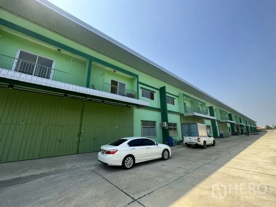 Warehouse for rent in Pathum Thani - Row of modern green warehouse units with wide driveway and parked vehicles.