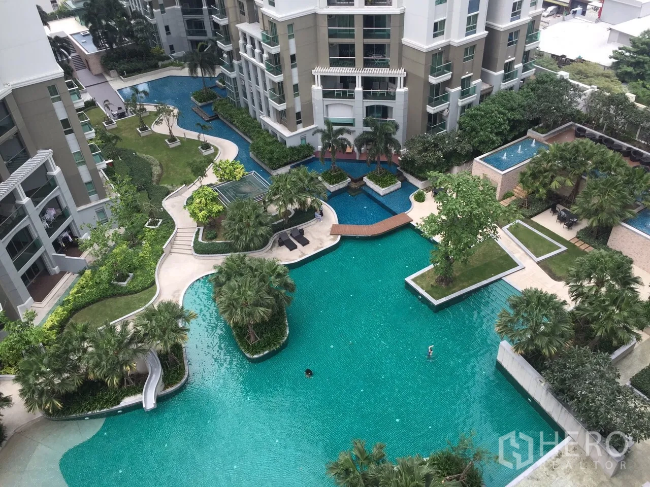 Condominium for rent in Huai Khwang, Bangkok - Aerial view of a resort-style lagoon pool with landscaped gardens, bridges and modern condominium towers.