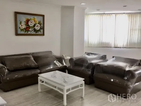 Condominium for rent in Watthana, Bangkok - Lounge area with brown sofas and armchairs in protective wrap, marble-top coffee table and large window with vertical blinds.
