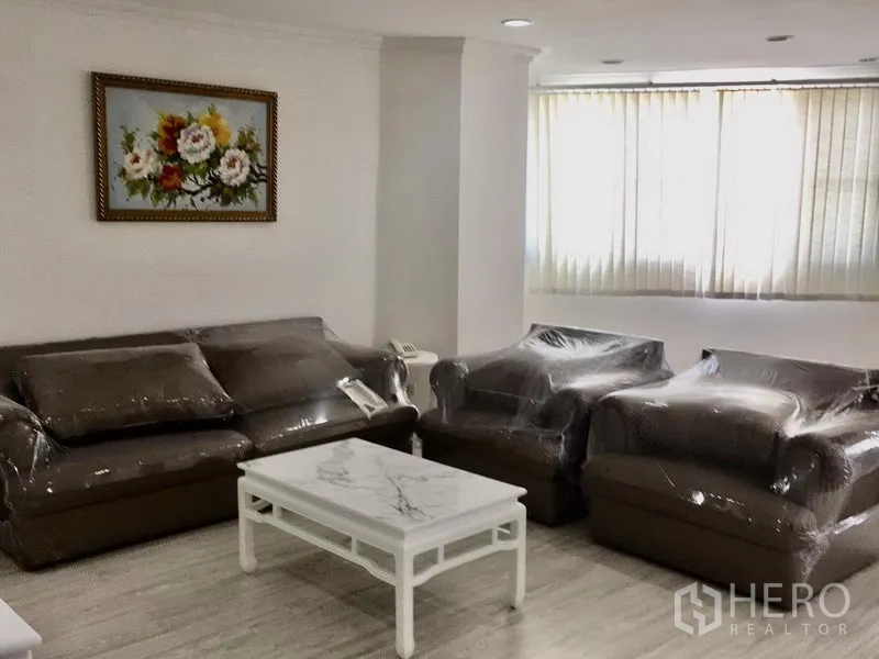 Condominium for rent in Watthana, Bangkok - Lounge area with brown sofas and armchairs in protective wrap, marble-top coffee table and large window with vertical blinds.