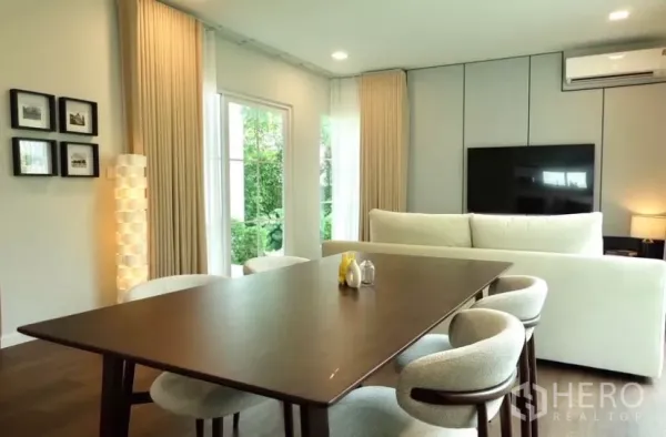 Detached House for rent in Bang Phli, Samut Prakan - Open-plan dining and living area with wood floors, large windows, beige curtains and a wall-mounted TV.