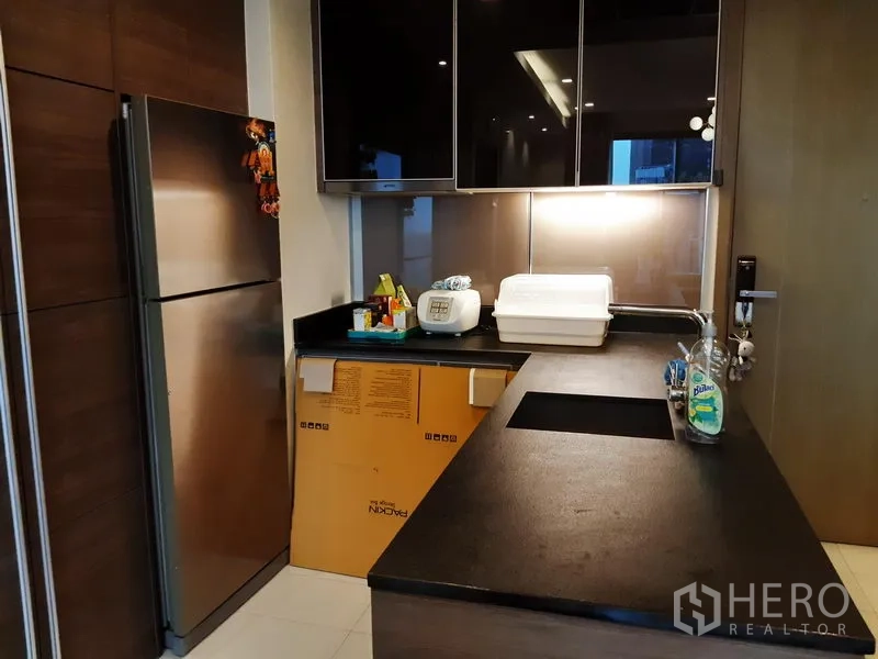 Condominium for rent in Khlong Toei, Bangkok - Compact modern kitchen with black stone countertop, under-mount sink, stainless refrigerator, and glossy upper cabinets.