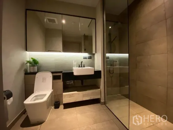 Condominium for rent or sale in Watthana, Bangkok - Modern bathroom with wall-mounted vanity, toilet, and a glass-enclosed rain shower with under-cabinet lighting.