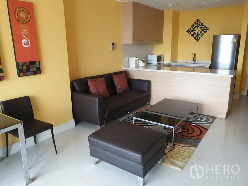 Condominium for rent or sale in Khlong Toei, Bangkok - Open-plan living area with brown sofa, ottoman, coffee table and a compact kitchenette with appliances.