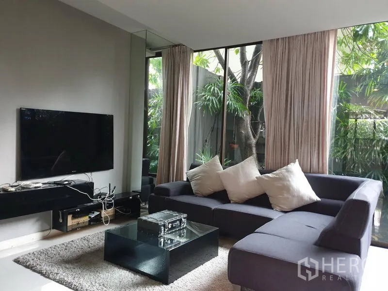 Detached House for sale in Watthana, Bangkok - Modern living room with L-shaped sofa, TV wall and floor-to-ceiling windows overlooking greenery.